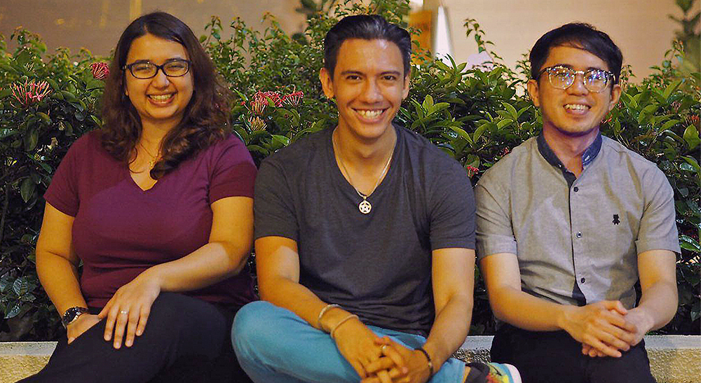 Three persons sitting on a ledge in front of some bushes, smiling into the camera.