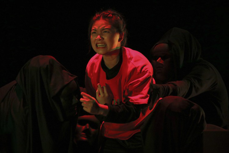 An angry-looking female-presenting person in a pink top, surrounded by people wearing black masks and black hoods.