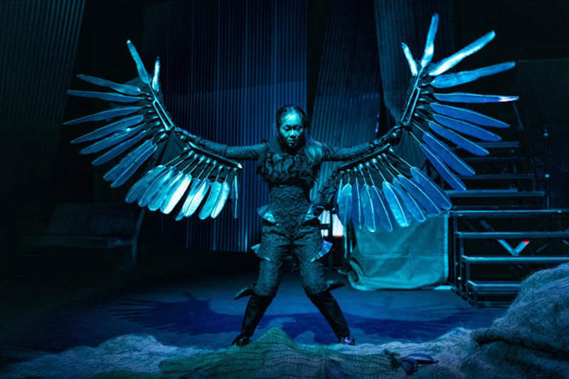 A person in a textured black full-body outfit, wearing large silver wings and tall black boots, standing on a dimly-lit stage.
