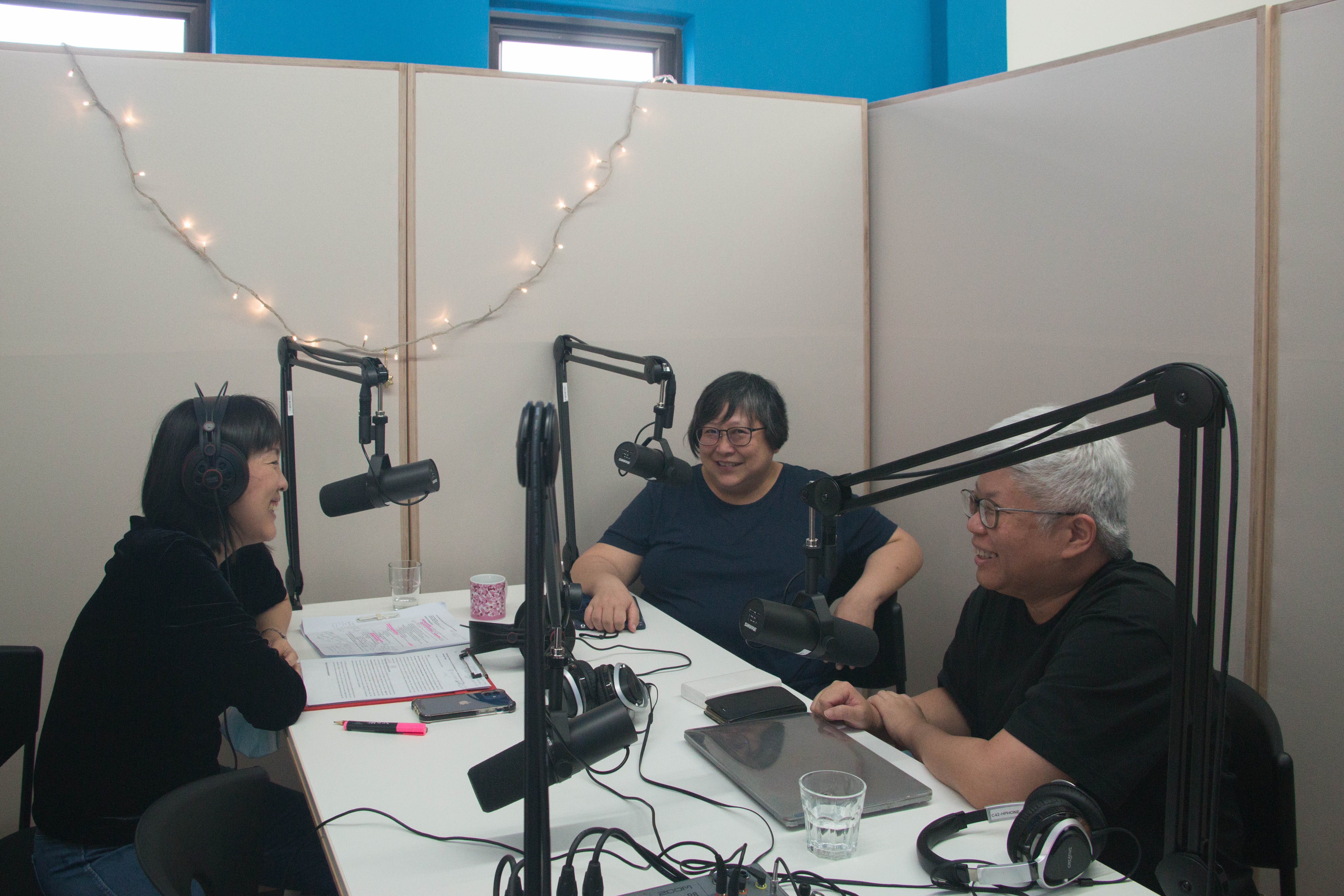 The photograph features a group of people in a conversation while seated around a table with microphones facing them, smiling as they wear headphones.