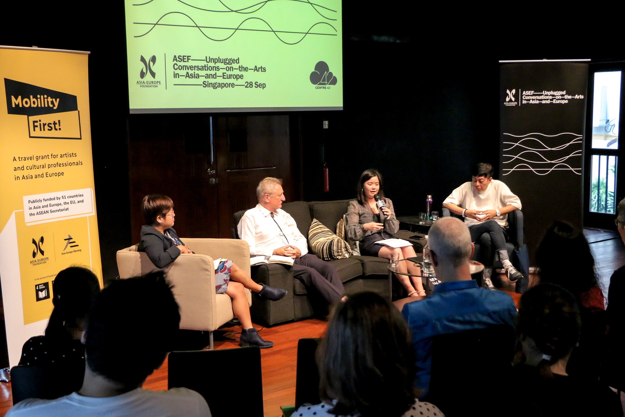 Photograph of ASEF Unplugged, with 4 speakers seated on couches at the Centre 42 Black Box. A projector screen is seen behind them.