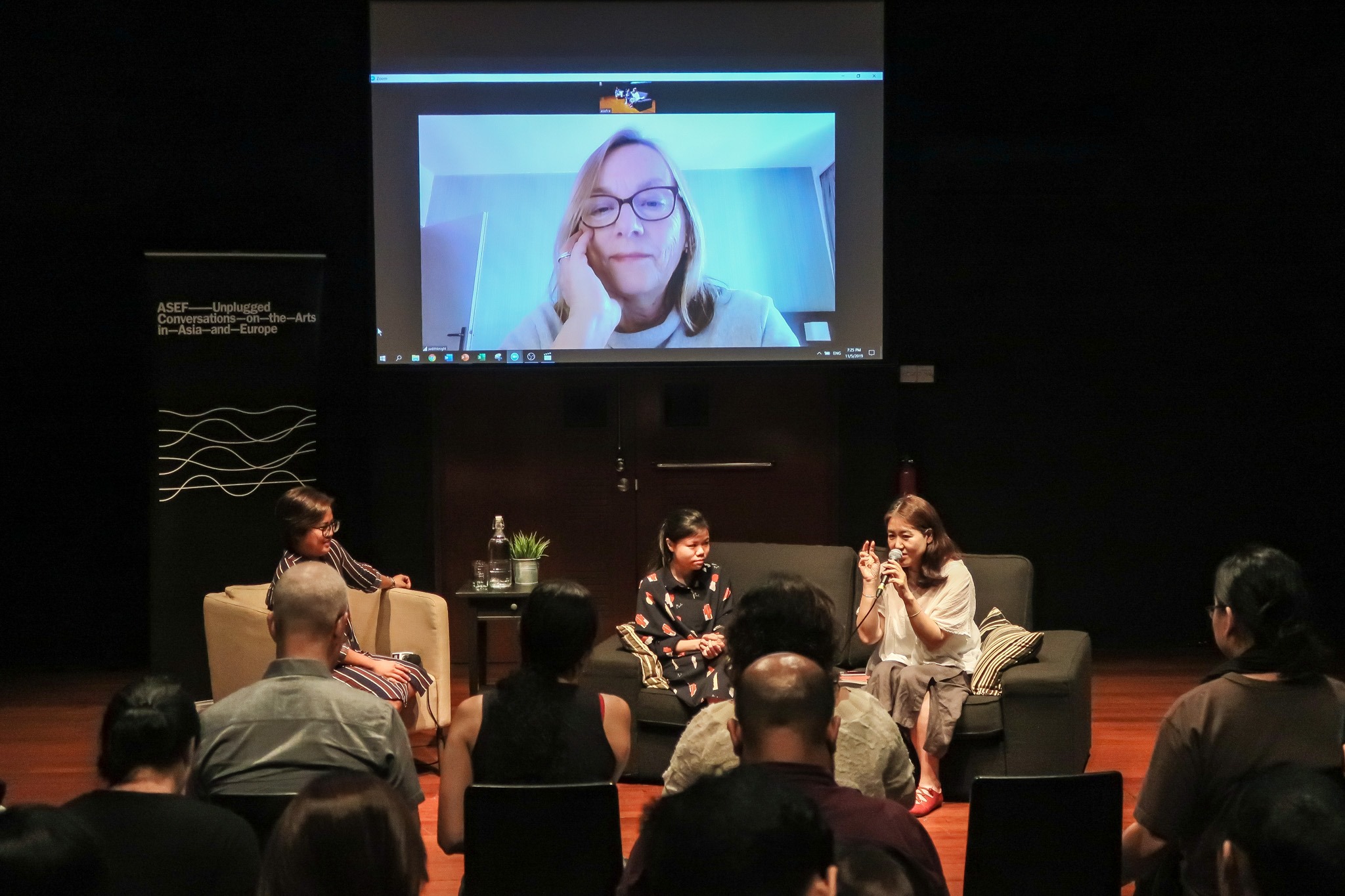 Photograph of ASEF Unplugged, with 3 speakers seated on couches at the Centre 42 Black Box. A projector screen is seen behind them.