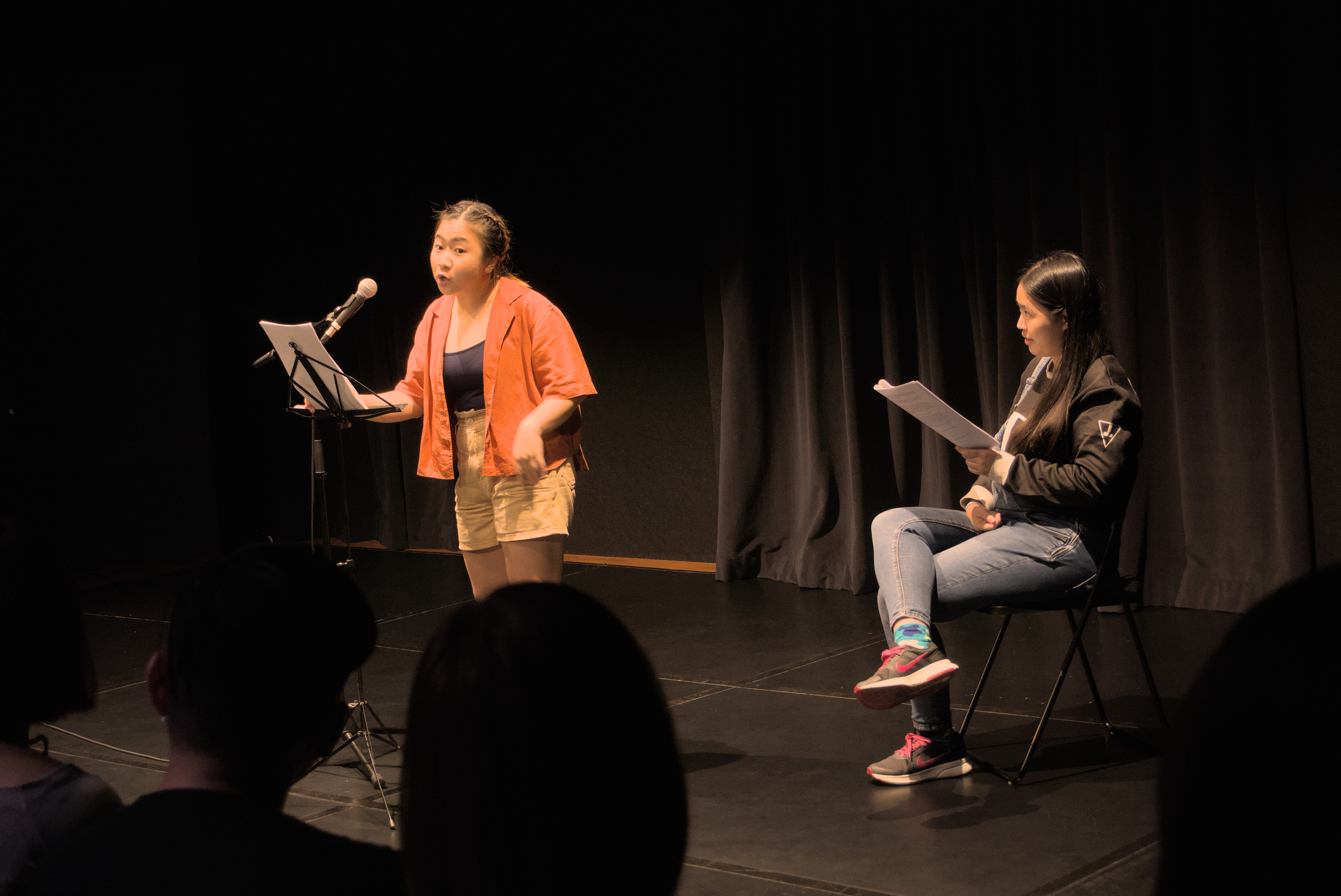 Cheryl Ho stands in front of a microphone and performs, while Cheryl Tan is seated next to her reading from a script.