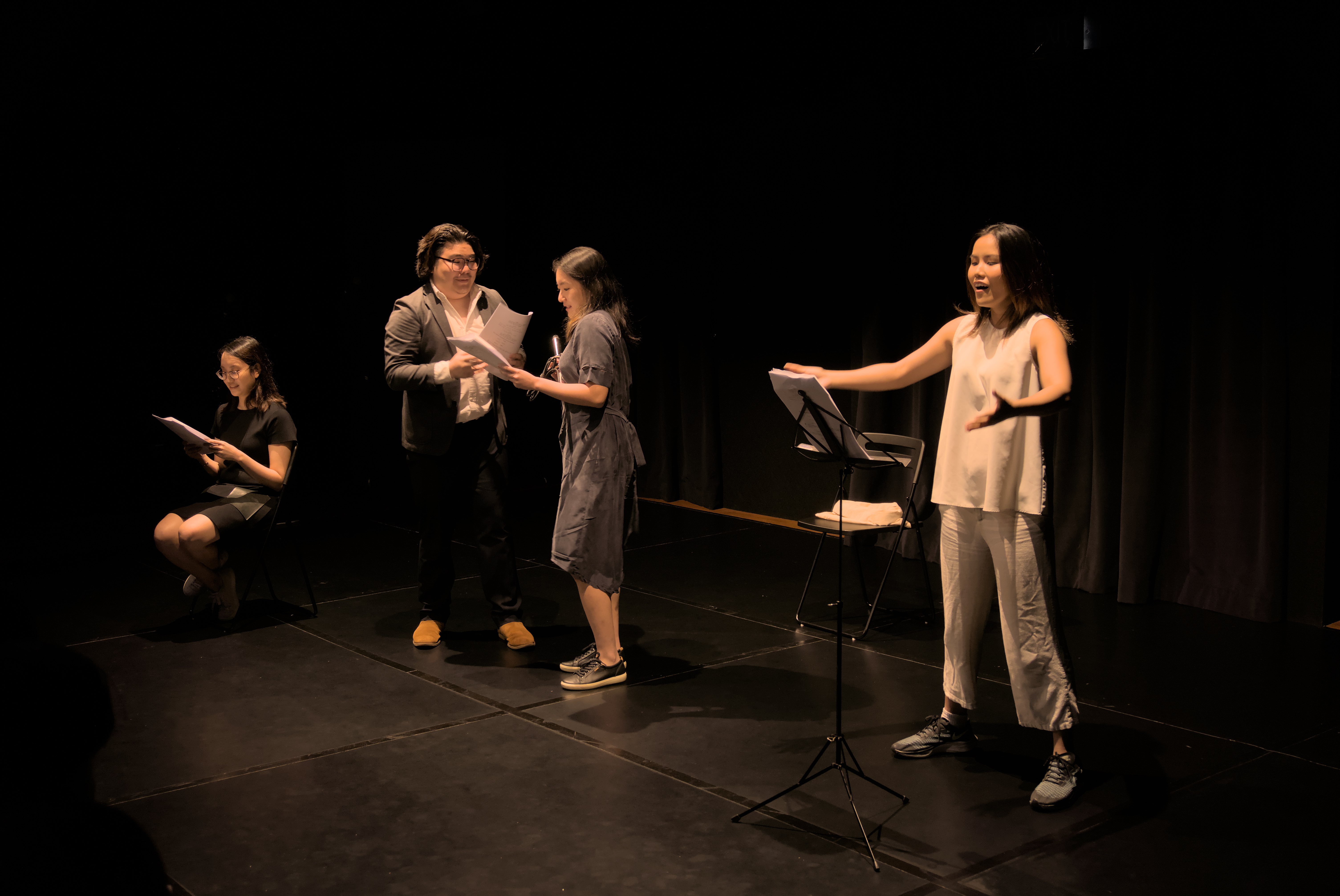 From left to right, Mary Tay is seated and reading from a script, Nicholas Chen is speaking to Claire Tay, and Rachel Chin is standing on the far end with her arms outstretched.